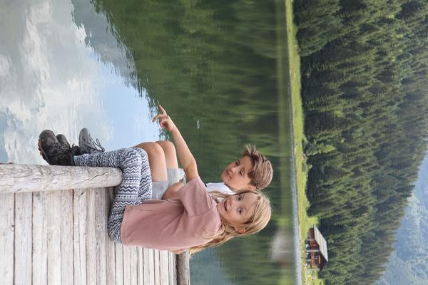 Two children are sitting on a wooden dock by a serene lake, surrounded by a lush green forest and mountains